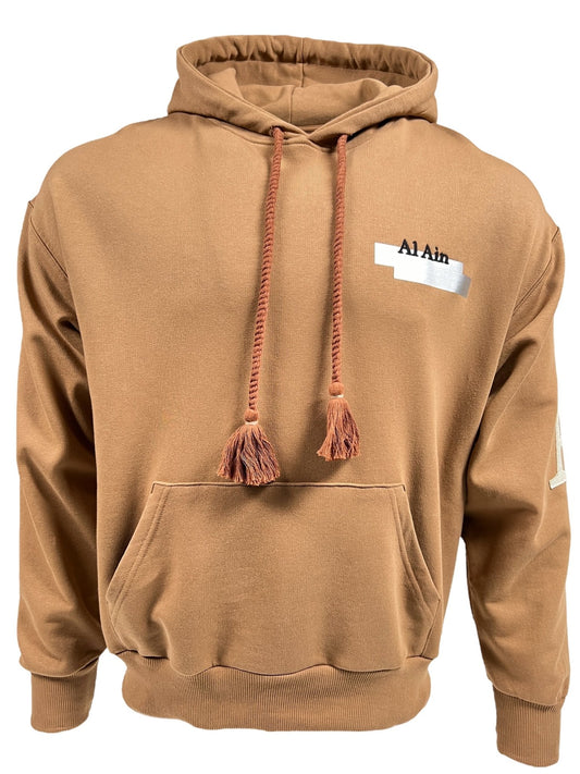 A brown AL AIN AHOX S102 PALMIER CHAMEAU hoodie with a front pocket and two drawstrings, featuring the text "Al Ain" on the left chest area and a stylish graphic back, perfect for streetwear enthusiasts.