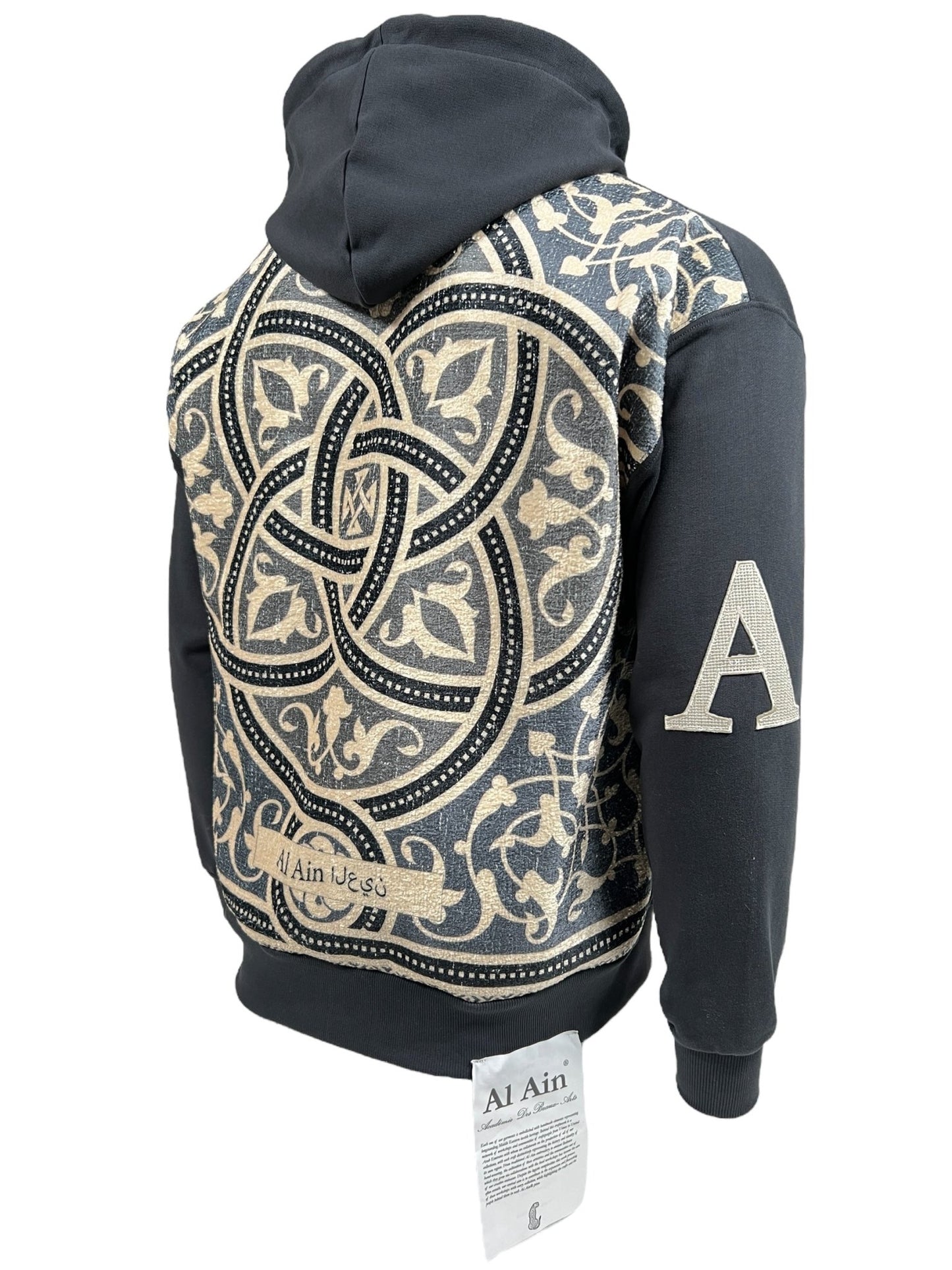 The AL AIN AHZX S103 SECRET NOIR hoodie, crafted from 100% cotton, showcases a black design with a complex beige and black pattern on the back. It features a large letter "A" on the left sleeve and a white label with text at the bottom, perfect for streetwear enthusiasts.