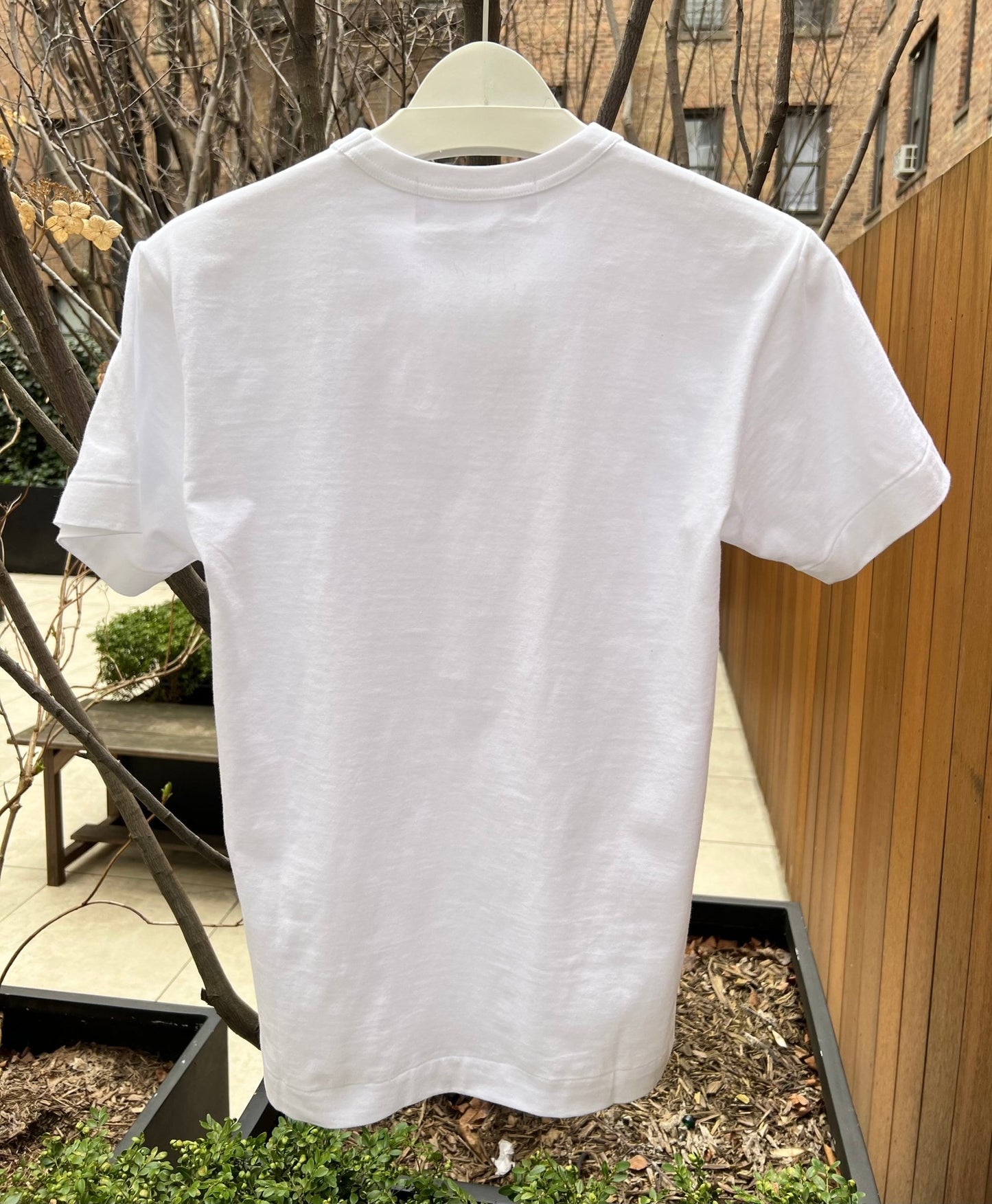 A COMME DES GARCONS COMMES DES GARCONS P1T064 PLAY T-SHIRT BLACK HEART BLACK made of 100% cotton is displayed on a hanger outdoors near potted plants and a wooden fence.