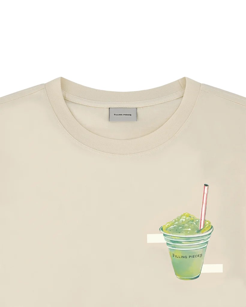 Close-up of a FILLING PIECES ICE VENDOR ANTIQUE WHITE T-SHIRT made of organic cotton with the label "filling pieces" and a graphic of a green drink with pearls, labeled similarly.