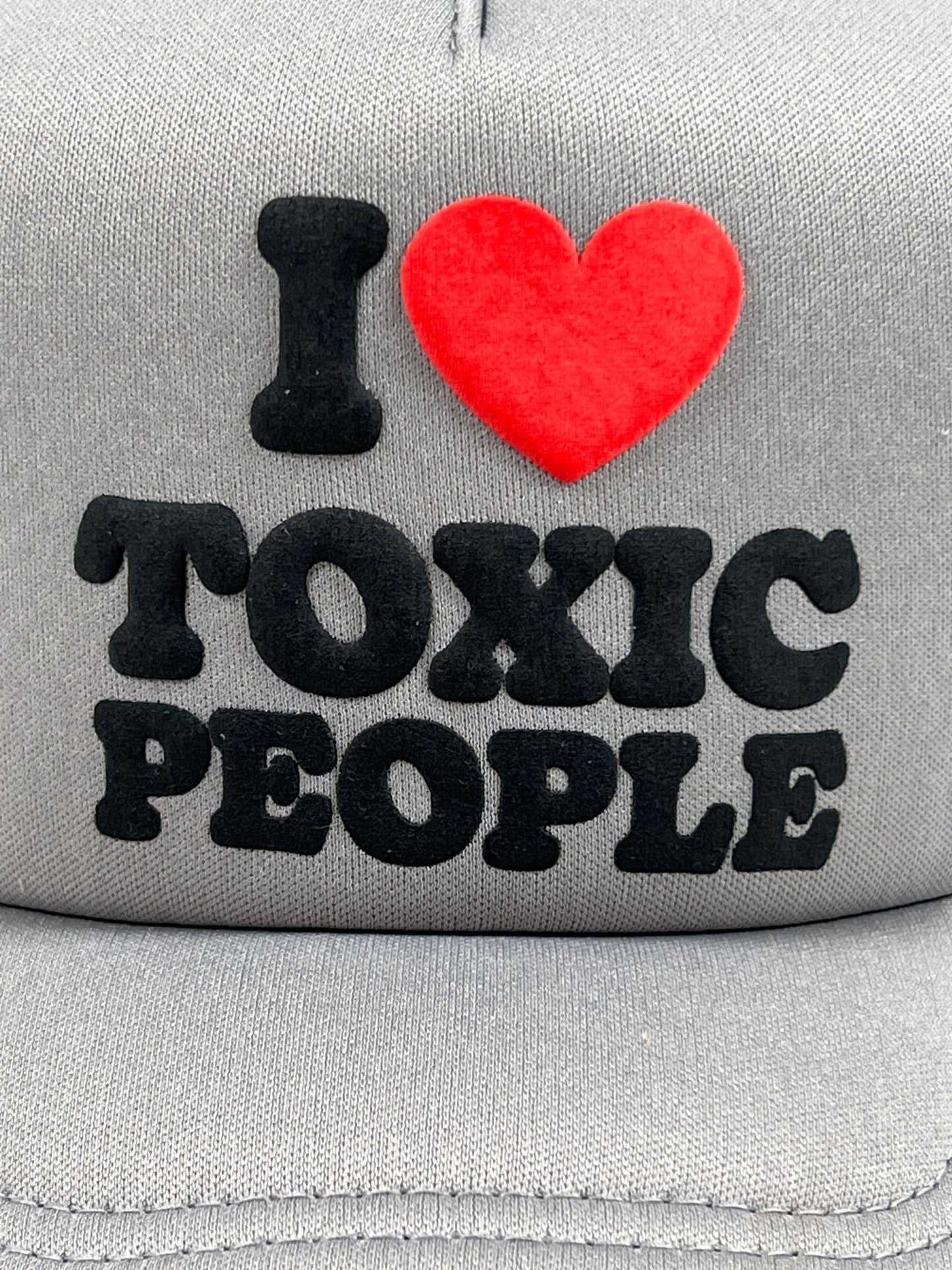A PLEASURES TOXIC TRUCKER CAP GREY, featuring the bold text "I ♥ TOXIC PEOPLE" in black and red letters, makes for a stylish accessory perfect for casual outings.