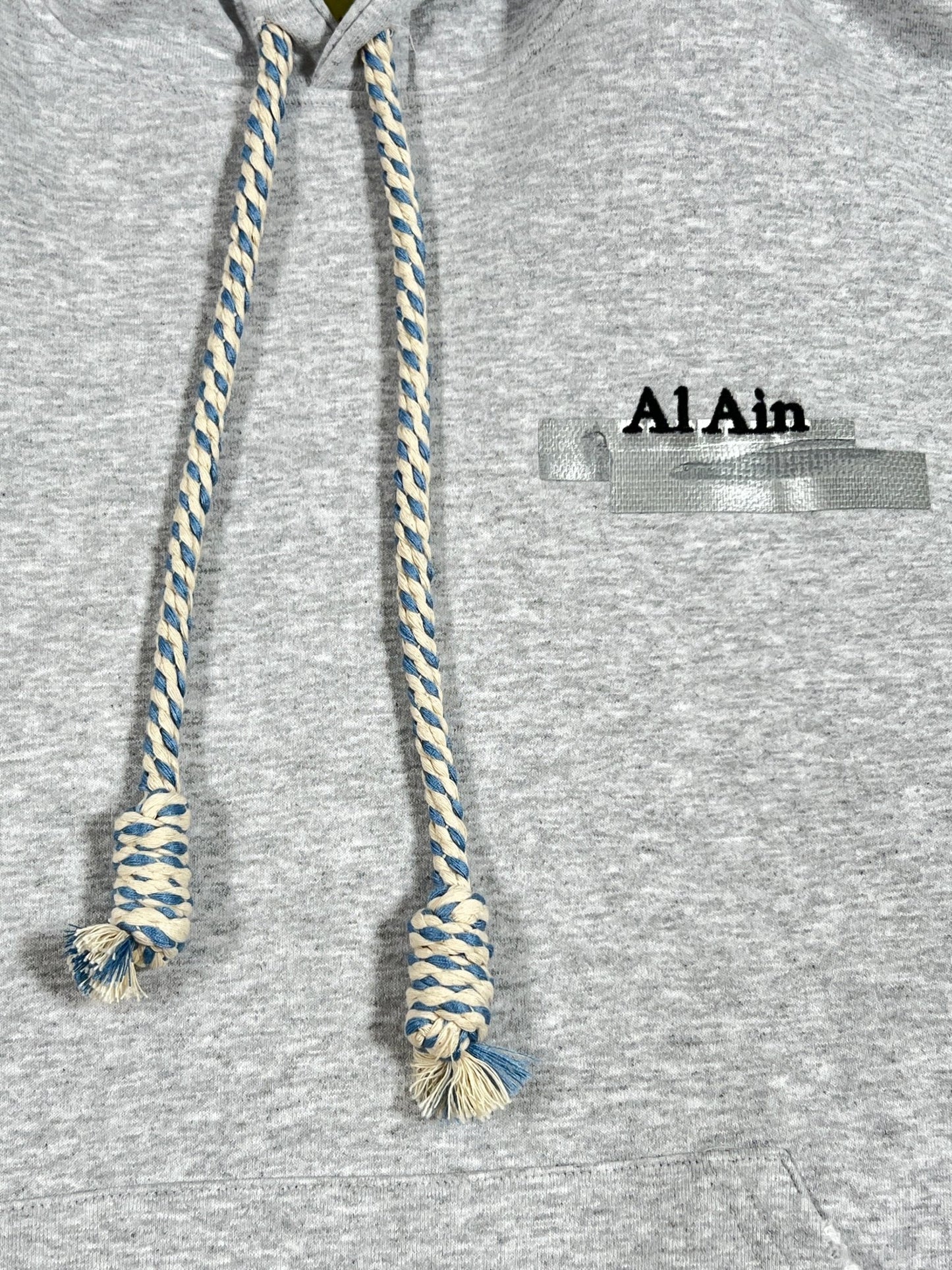 Close-up of a grey AL AIN AHOX S150 YACHT LIMITED hoodie with blue and white braided drawstrings. The text "AL AIN" is visible on the chest, partially covered by tape. Thick rope texture lace detail enhances the hoodie, giving it an extra touch of style.