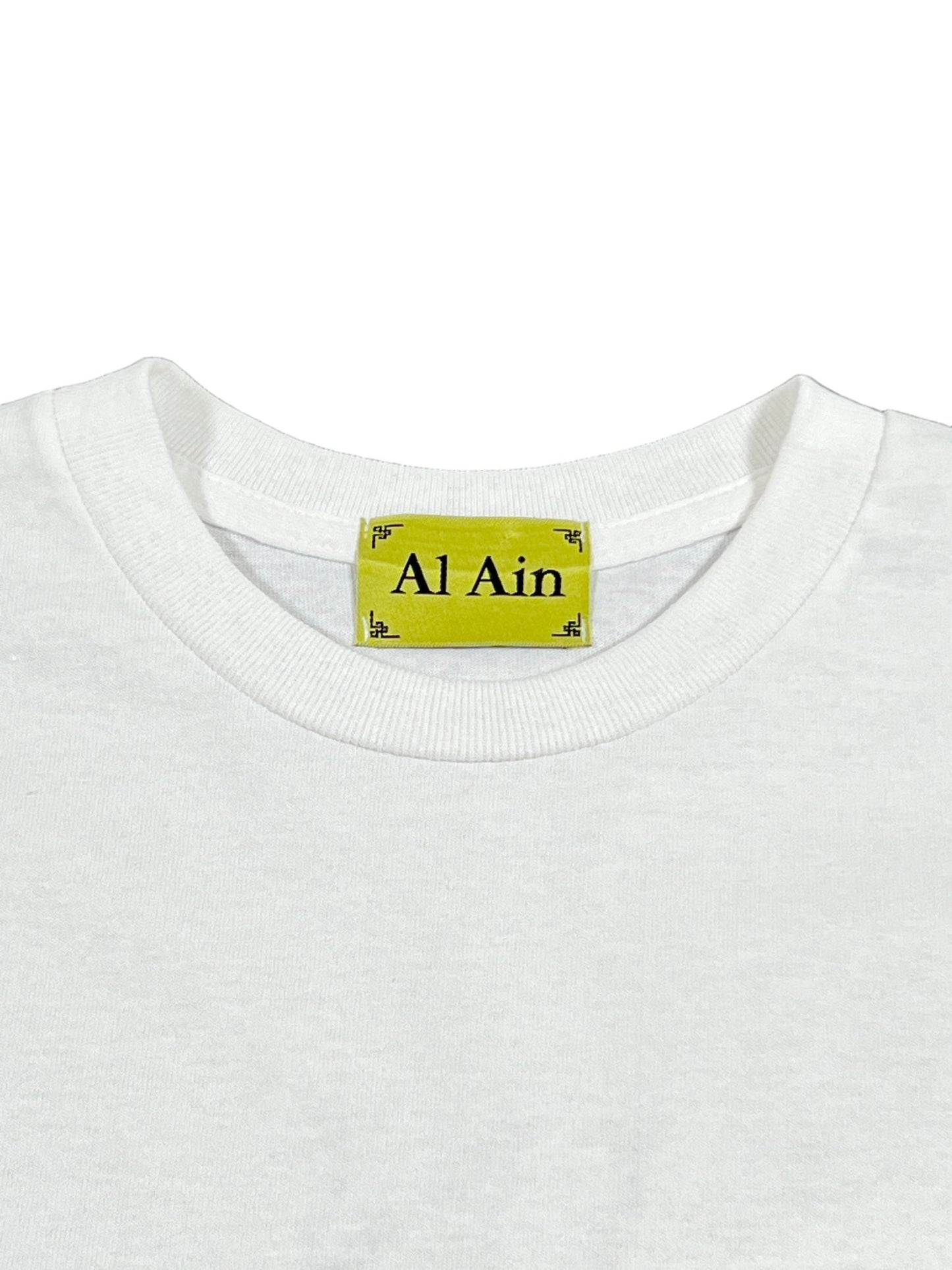 White T-shirt with a large graphic detail featuring a green label displaying the product name "AL AIN AMHX S121 PEACOCK BLANC" and brand name "AL AIN.