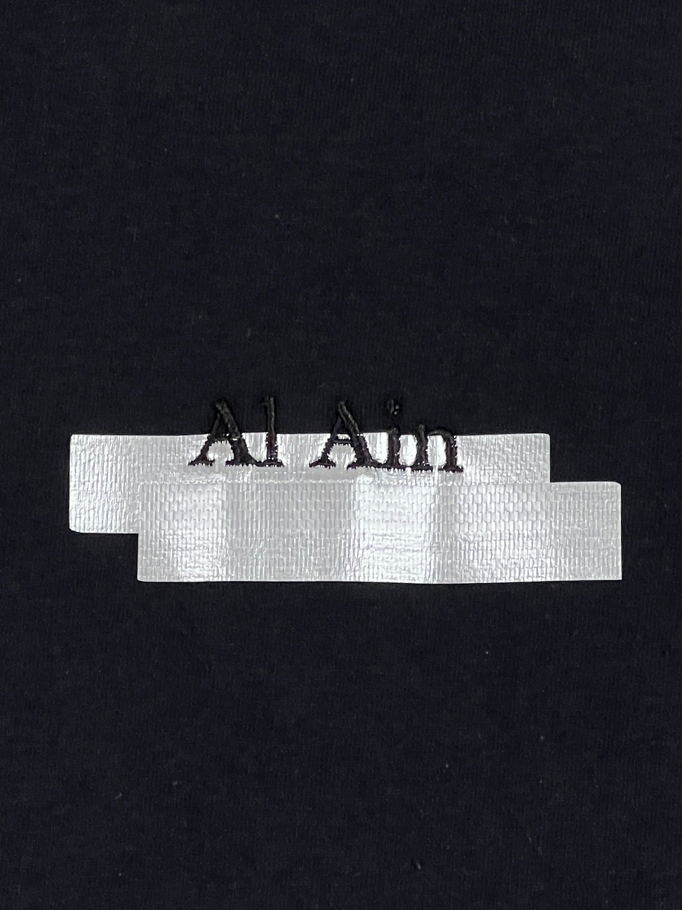 A piece of black fabric with the word "AL AIN AMHX S124 CHILL NOIR" embroidered in black thread, partially covered by horizontal strips of silver ribbon—creating a stylish and eye-catching design reminiscent of a graphic t-shirt.
