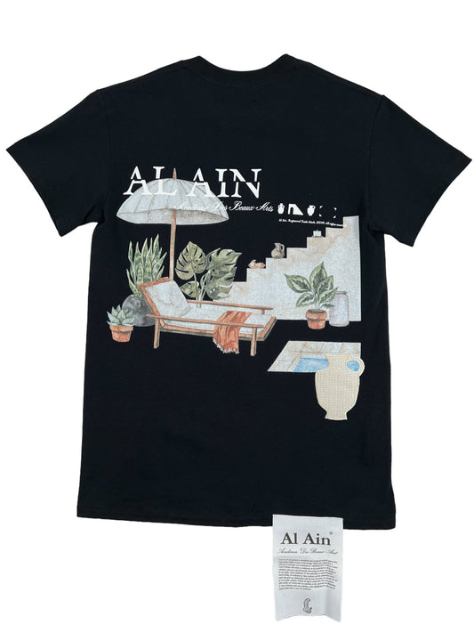This stylish and eye-catching black graphic t-shirt features a poolside scene with plants, a lounge chair, and an umbrella. The text "Al Ain" is embroidered at the top. A small tag is displayed in front of the shirt. Introducing the AL AIN AMHX S124 CHILL NOIR by AL AIN.