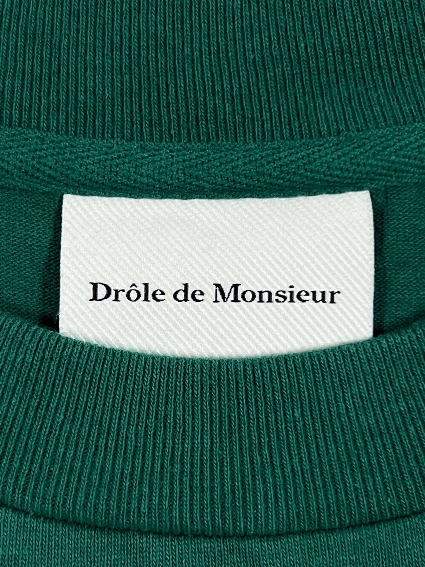 Close-up of a clothing label reading "DROLE DE MONSIEUR" on a white background sewn into a green ribbed neckline.