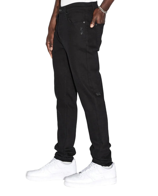 A man wearing KSUBI black denim jeans and white sneakers.