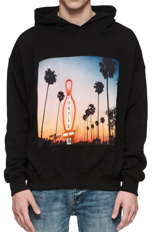 A man wearing a PURPLE BRAND P494-PBCS PURPLE X BLUE SKY HOODY BLACK with a graphic image of palm trees.