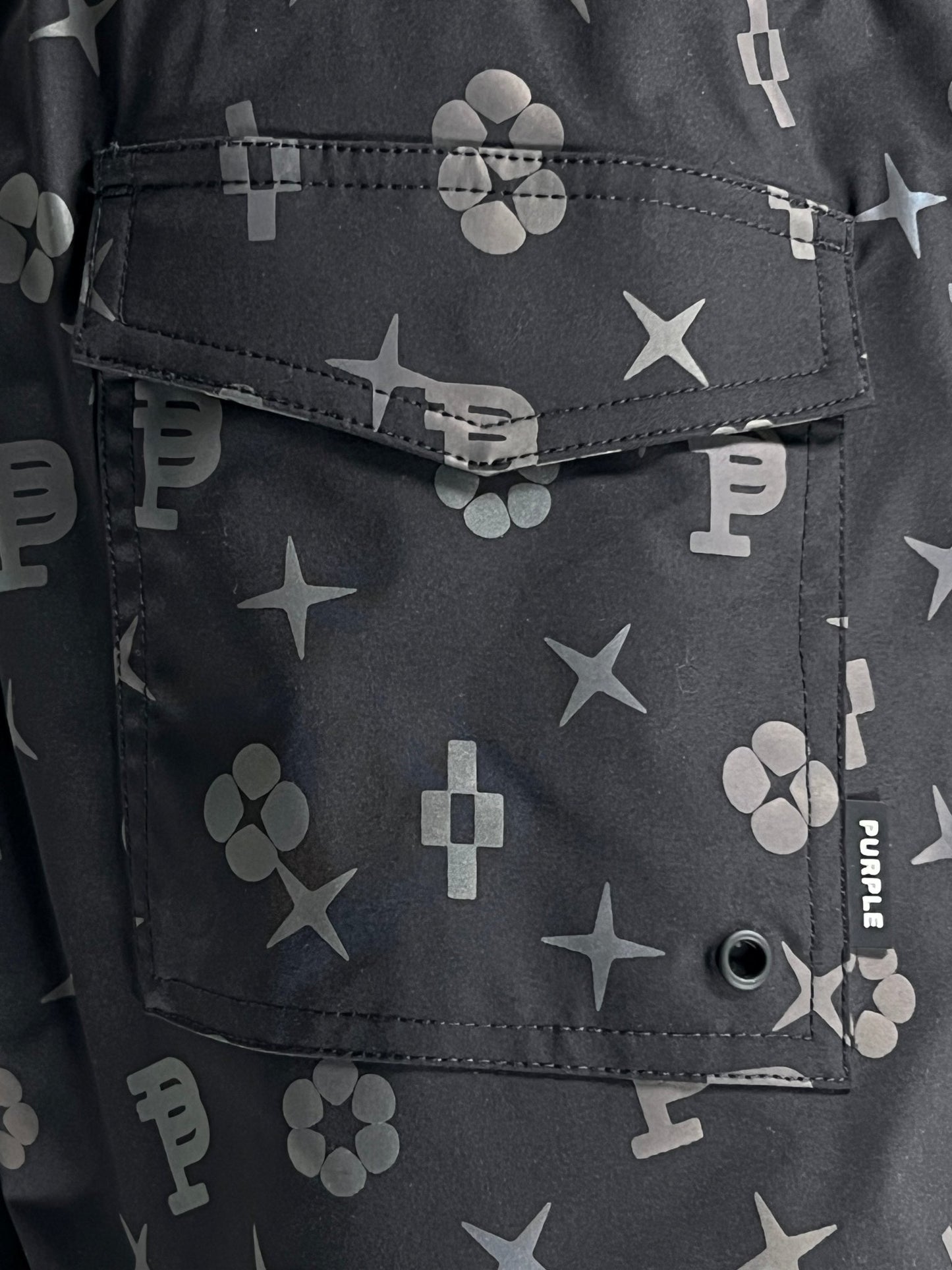 Close-up of a black fabric pocket with various gray symbols, including stars, crosses, and letters, along with the word "PURPLE." The pocket has a flap secured with a buttonhole, highlighting the unique design often seen in PURPLE BRAND P504-PBBH ALL ROUND SHORT AOP by PURPLE BRAND.