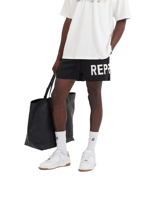 A man in a white shirt and REPRESENT SWIM SHORTS BLK holding a black tote bag.