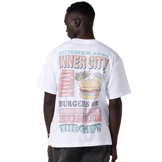 A man showcases a retro vibe with the Honor The Gift Htg Burgers Ss Tee in white, crafted from 100% cotton and garment dyed. This vintage pocket T-shirt features vibrant graphic text and images of a burger, along with the phrases "Inner City Summer 2023" and "Honor the Gift," enhancing its nostalgic appeal.