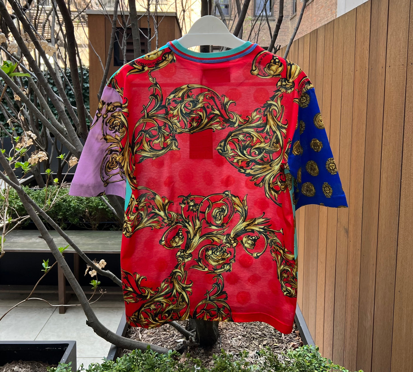 A colorfully patterned VERSACE E72GAH6O9-EJS064-EG12 T-SHIRT displayed outdoors on a clear hanger against a backdrop of plants and wooden fencing.