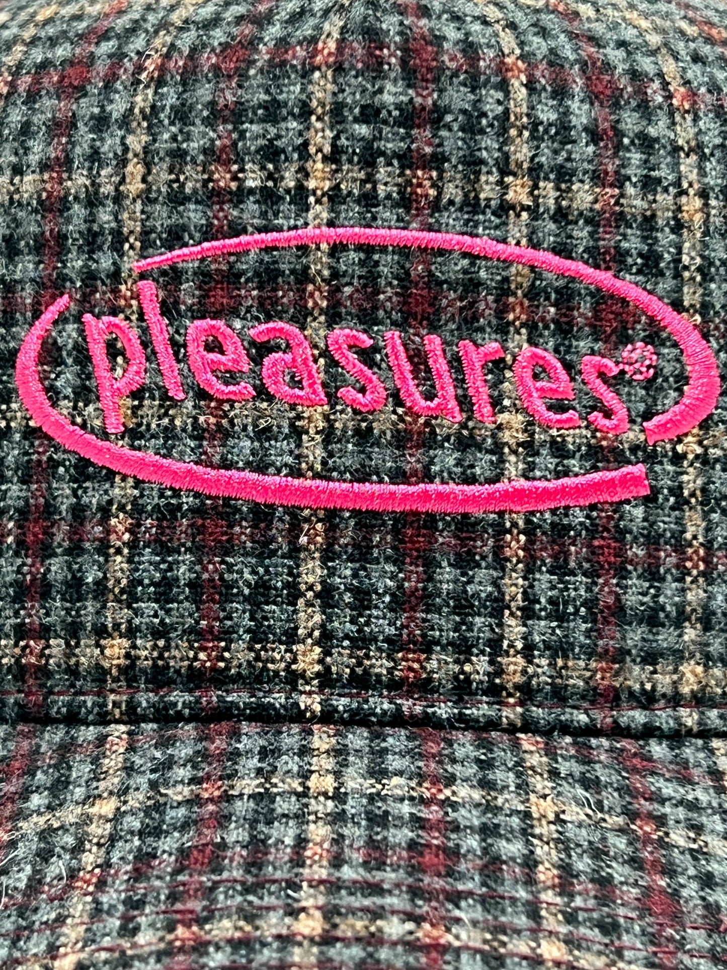 Close-up of a checkered fabric PLEASURES HAPPIER PLAID TRUCKER MAROON hat, with the word "pleasures" embroidered in pink within an oval outline.