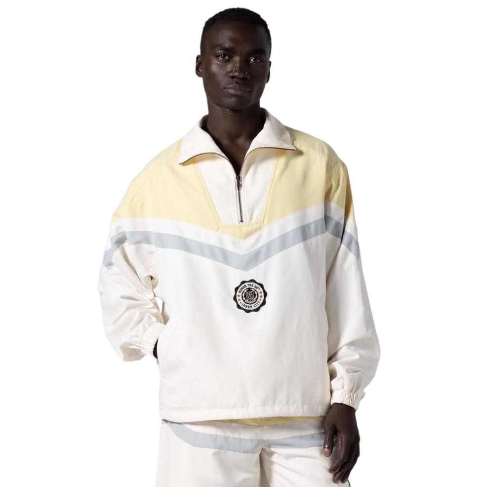 A fashion-forward individual sports the Honor The Gift A-Spring Brushed Poly Track Anorak Bone by HONOR THE GIFT, showcasing a white and yellow zip-up design with gray stripes and a circular logo on the front, crafted from durable materials.