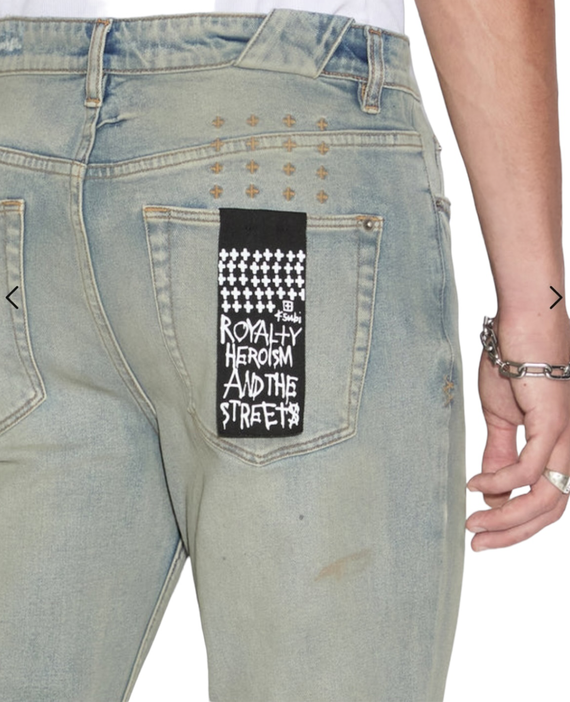 Back view of a person wearing KSUBI CHITCH MECHANIK DENIM jeans in light blue, with a black and white patch on the right back pocket displaying the text "Royalty Heroism And The Streets." The person complements their outfit with a silver bracelet.