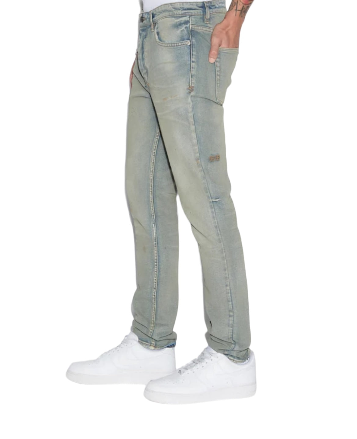 A person with visible tattoos, wearing KSUBI's CHITCH MECHANIK DENIM—slim tapered jeans made from stretch denim—and white sneakers stands against a plain background.