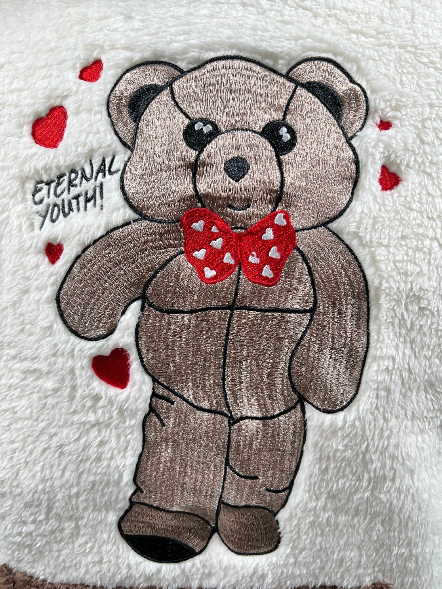 The Advisory Board Crystals Bear Patch Boa Fleece Hoodie in ecru boasts a charming teddy bear patch adorned with a red bow tie and hearts on its cozy fabric. The phrase "ETERNAL YOUTH" is creatively embroidered next to the patch, embodying the whimsical essence of ADVISORY BOARD CRYSTALS.