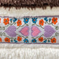 A close-up of a stylish fleece fabric reveals embroidered pink hearts along with orange and blue flowers, all bordered by soft, textured white and brown materials, reminiscent of the ADVISORY BOARD CRYSTALS Bear Patch Boa Fleece Hoodie Ecru.