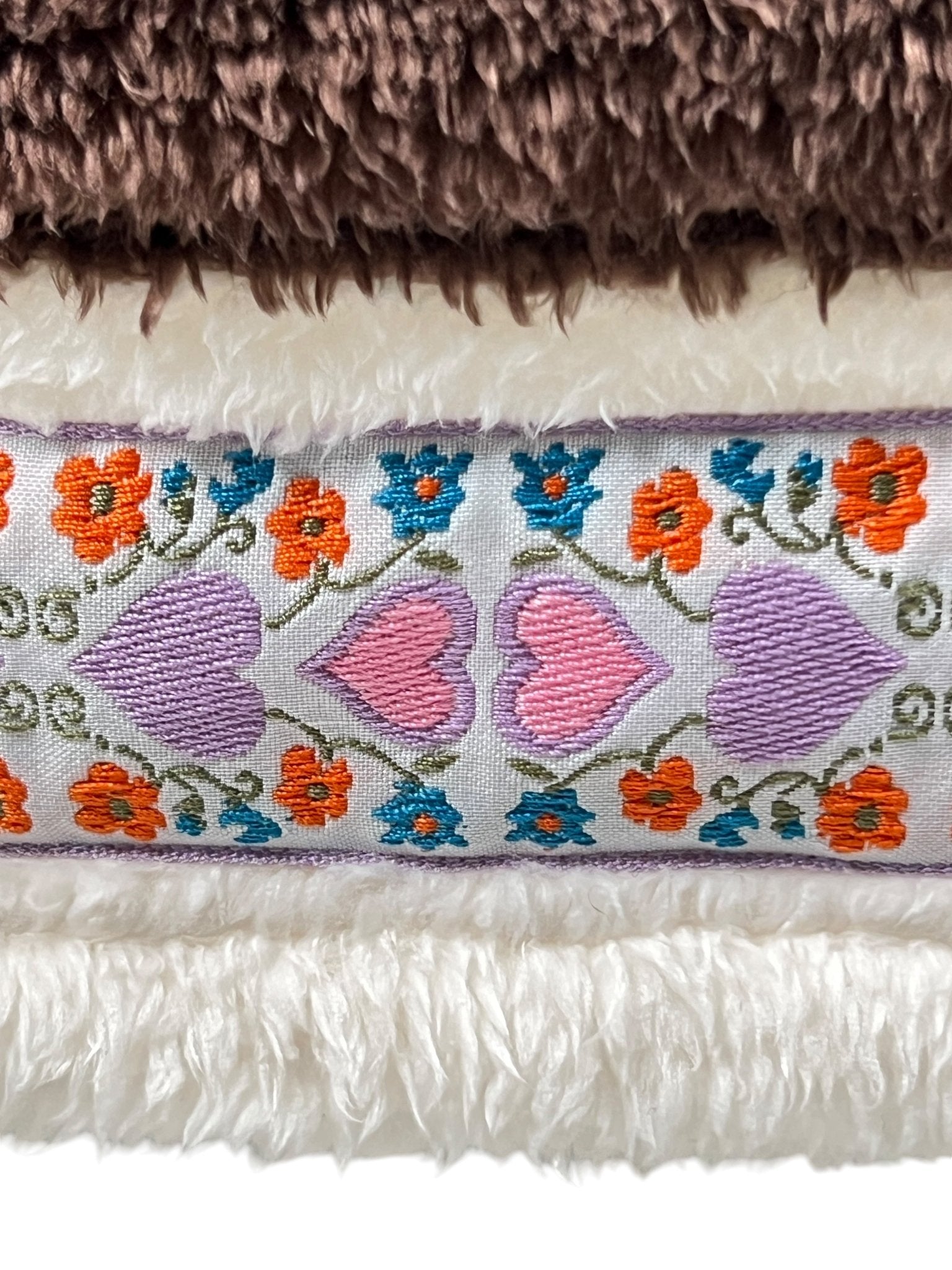 A close-up of a stylish fleece fabric reveals embroidered pink hearts along with orange and blue flowers, all bordered by soft, textured white and brown materials, reminiscent of the ADVISORY BOARD CRYSTALS Bear Patch Boa Fleece Hoodie Ecru.