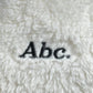 Close-up of the word "Abc." embroidered in black thread on a fluffy white fabric, reminiscent of the stylish fleece found on an Ecru Bear Patch Boa Fleece Hoodie by ADVISORY BOARD CRYSTALS.