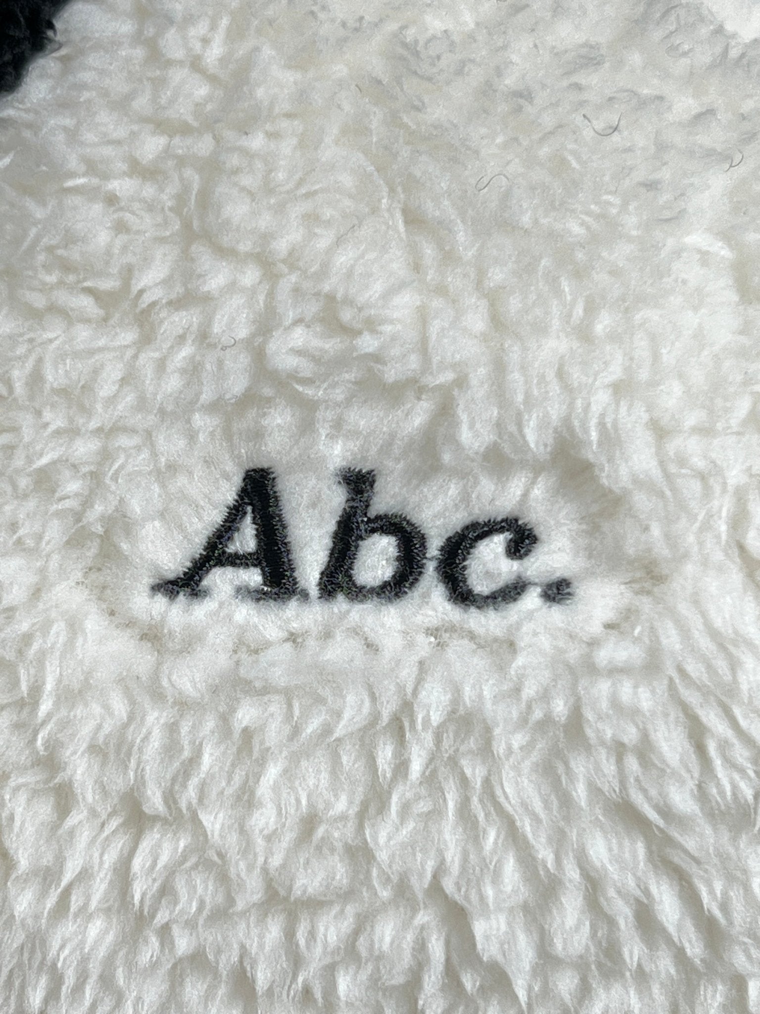 Close-up of the word "Abc." embroidered in black thread on a fluffy white fabric, reminiscent of the stylish fleece found on an Ecru Bear Patch Boa Fleece Hoodie by ADVISORY BOARD CRYSTALS.