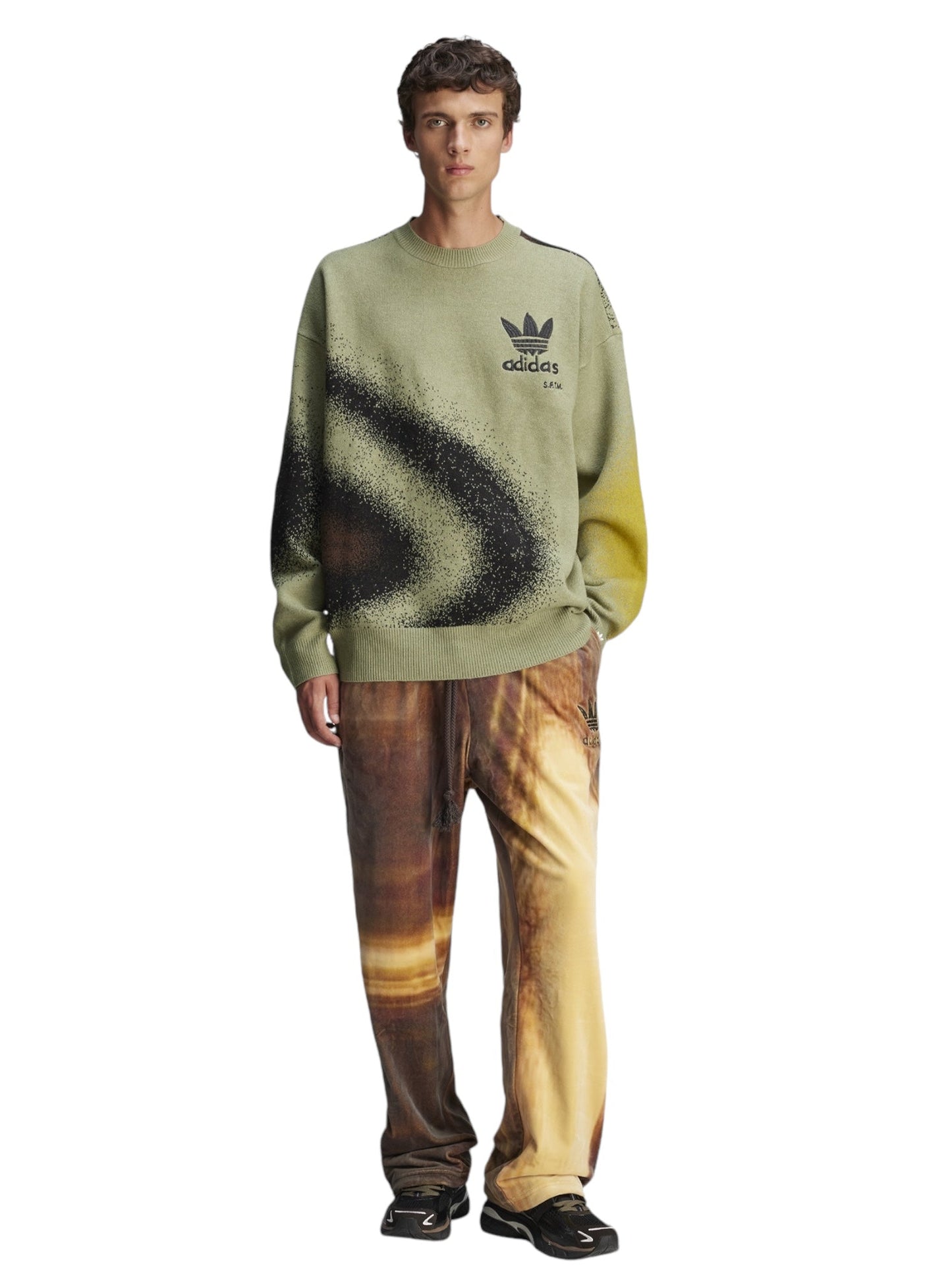 A person wearing the Adidas X Song For The Mute Ji9643 Adi004 Knitswea Tengr in a green color, adorned with a black graphic, and paired with multicolored pants stands against a white background, reflecting the distinctive style of ADIDAS x SONG FOR THE MUTE.