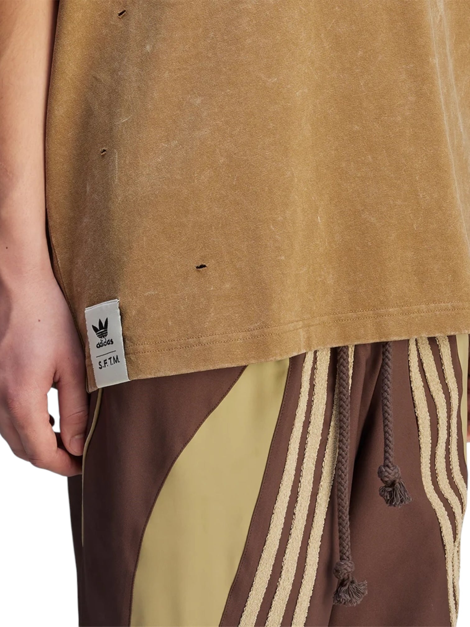 A person wears the Adidas x Song For The Mute KA7995 SFTM Wash Tee in EARTH with brown track pants featuring beige stripes and drawstrings from ADIDAS x SONG FOR THE MUTE.