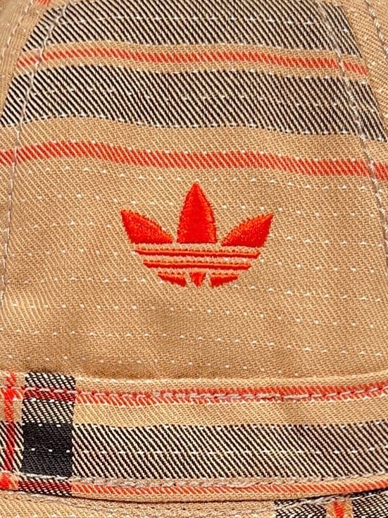 Close-up of a textured 100% cotton fabric with a stitched red ADIDAS X WALES BONNER IW1163 WB HAT BEIGWB- logo on it.