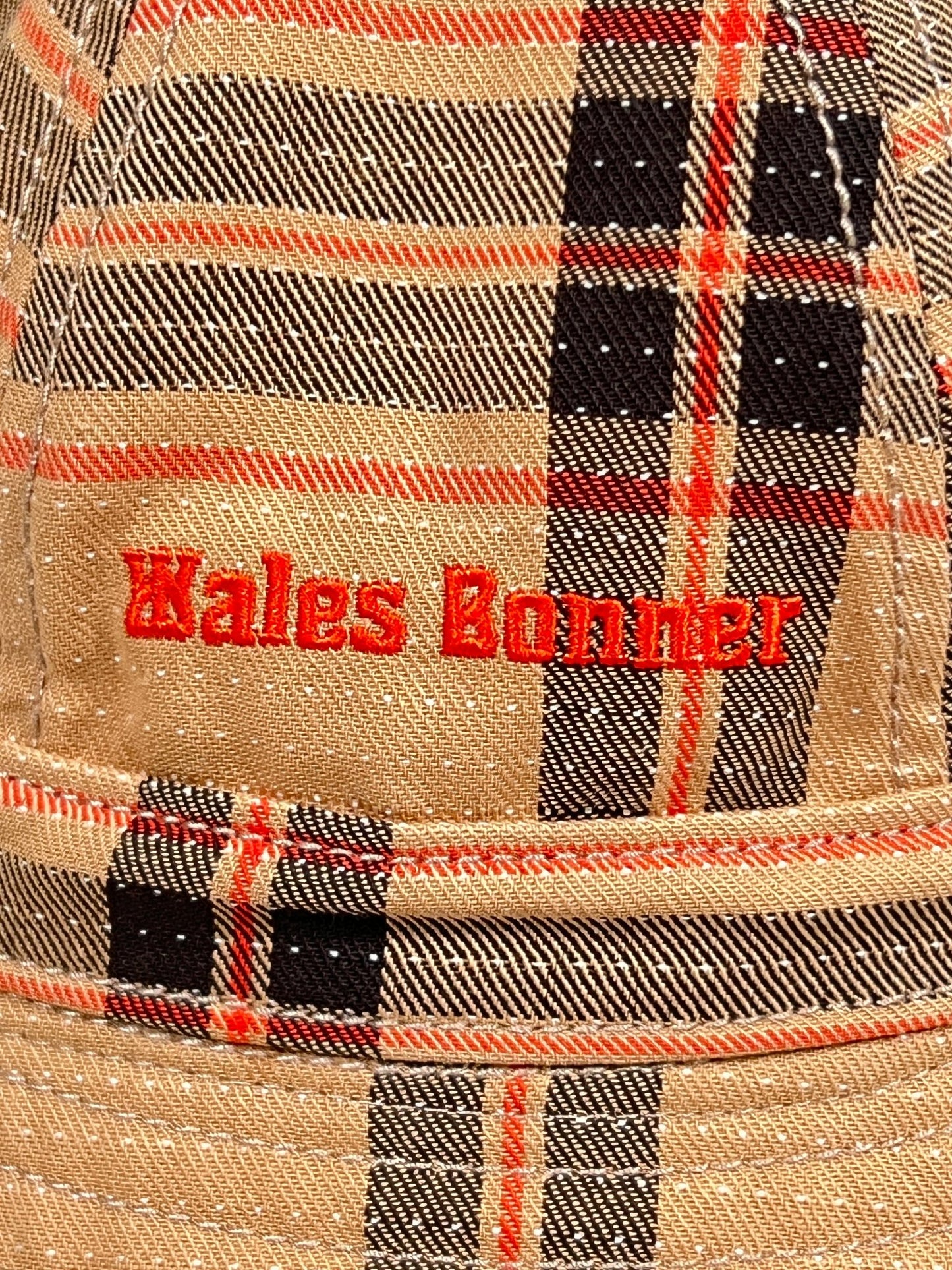 Close-up of fabric with the red embroidered text "ADIDAS X WALES BONNER IW1163 WB HAT BEIGWB-" on a beige plaid patterned background with black, red, and white lines.