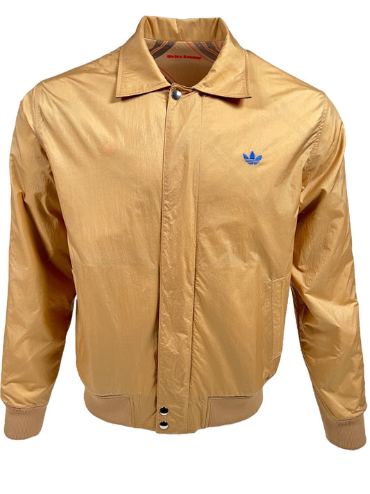 A golden ADIDAS x WALES BONNER bomber jacket displayed on a white background, featuring a blue logo print on the left chest area and a two-way zip fastening.