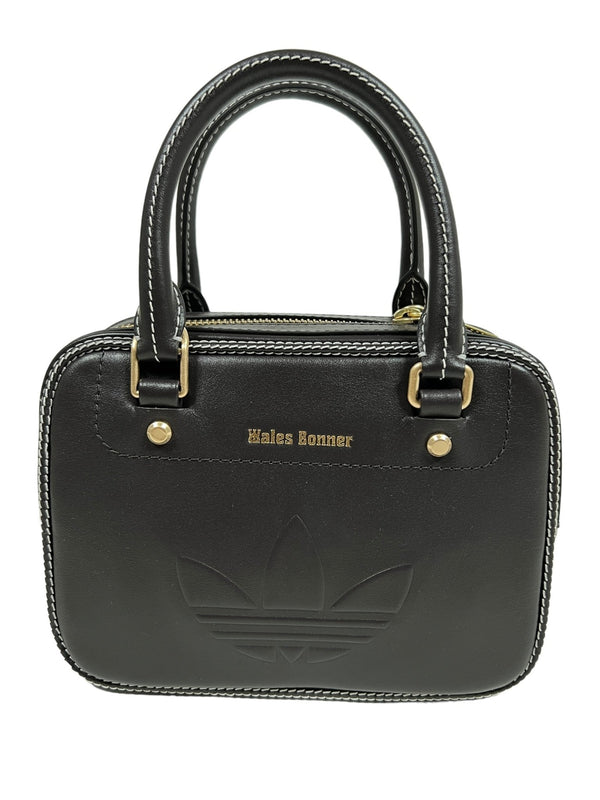 A small black leather handbag with double handles features white stitching, a gold "ADIDAS x WALES BONNER" logo, and an embossed design, capturing the timeless elegance of the collaboration.