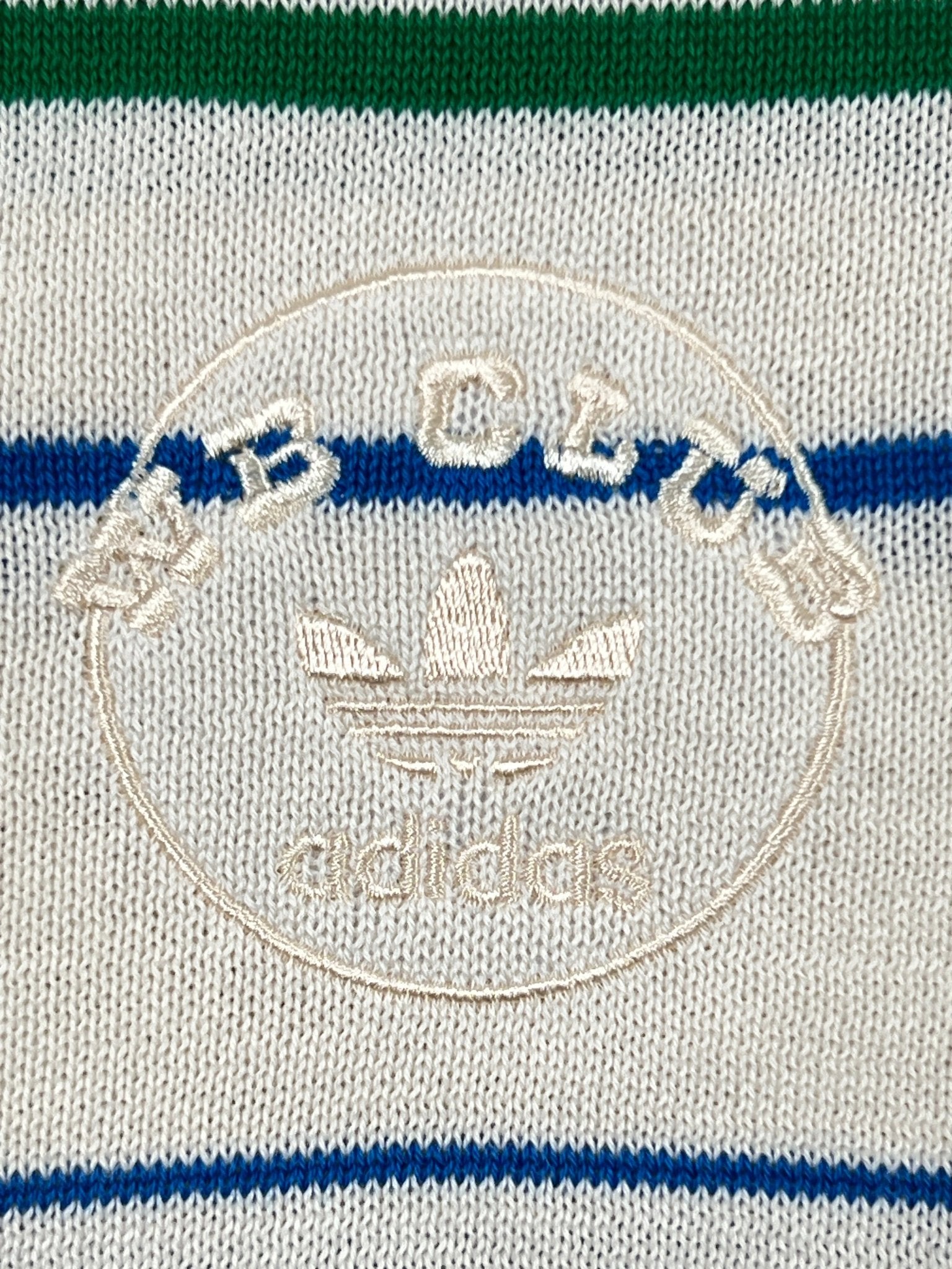 Close-up of the Adidas x Wales Bonner JM8203 WB Knit Polo WonWh, showcasing green and blue horizontal stripes and a beige embroidered circular logo—a vintage-inspired piece from ADIDAS x WALES BONNER.