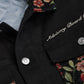 The Advisory Board Crystals Abc. Floral Jacquard & Canvas Jacket in black by ADVISORY BOARD CRYSTALS features floral embroidery on the collar and pockets, adorned with silver buttons and "Advisory Board" text on the chest. It is equipped with a quilted signature lining for enhanced comfort, making it a stylish and cozy choice.