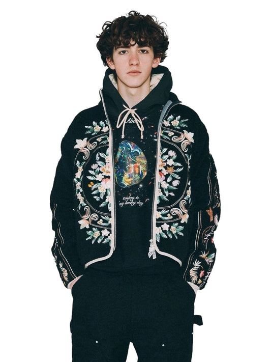 A person wearing an ADVISORY BOARD CRYSTALS Floral Fleece Zip-Up Black, adorned with vibrant floral embroidery, stands with hands in pockets against a plain background.