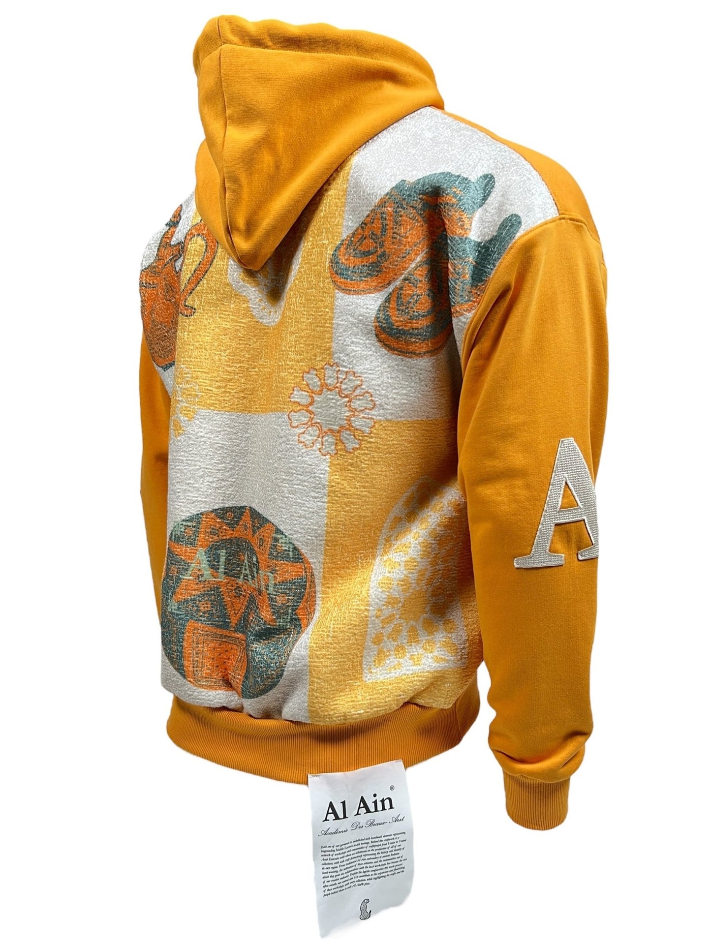 The Al Ain AHOX S108 Pouvoir Ocre by AL AIN is an ocre yellow hoodie adorned with colorful abstract patterns on the back. It features a stylish "A" on the sleeve and a label with text at the hem. Made from 100% cotton, this streetwear piece seamlessly blends comfort and style.