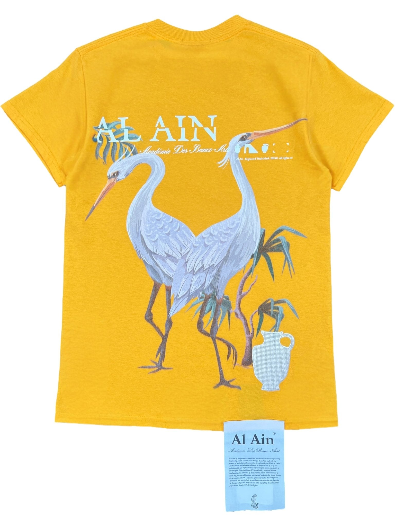 The Al Ain AMHX S120 Jabalia Ocre Yellow t-shirt features two white cranes with green plants and a white jug on the back, "AL AIN" in bold white text above, embroidered branding, and a blue "Al Ain" tag below.