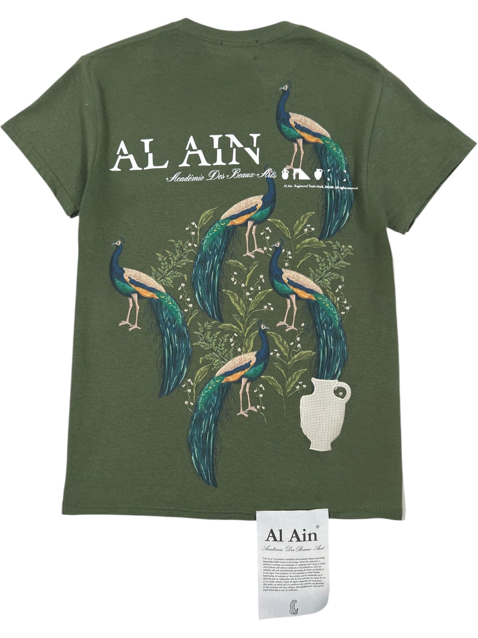The Al Ain AMHX S121 Peacock Militaire graphic t-shirt by AL AIN features bold “AL AIN” text, white script, six peacocks on branches above a white vase, a matching bottom tag, and an embroidered accent.