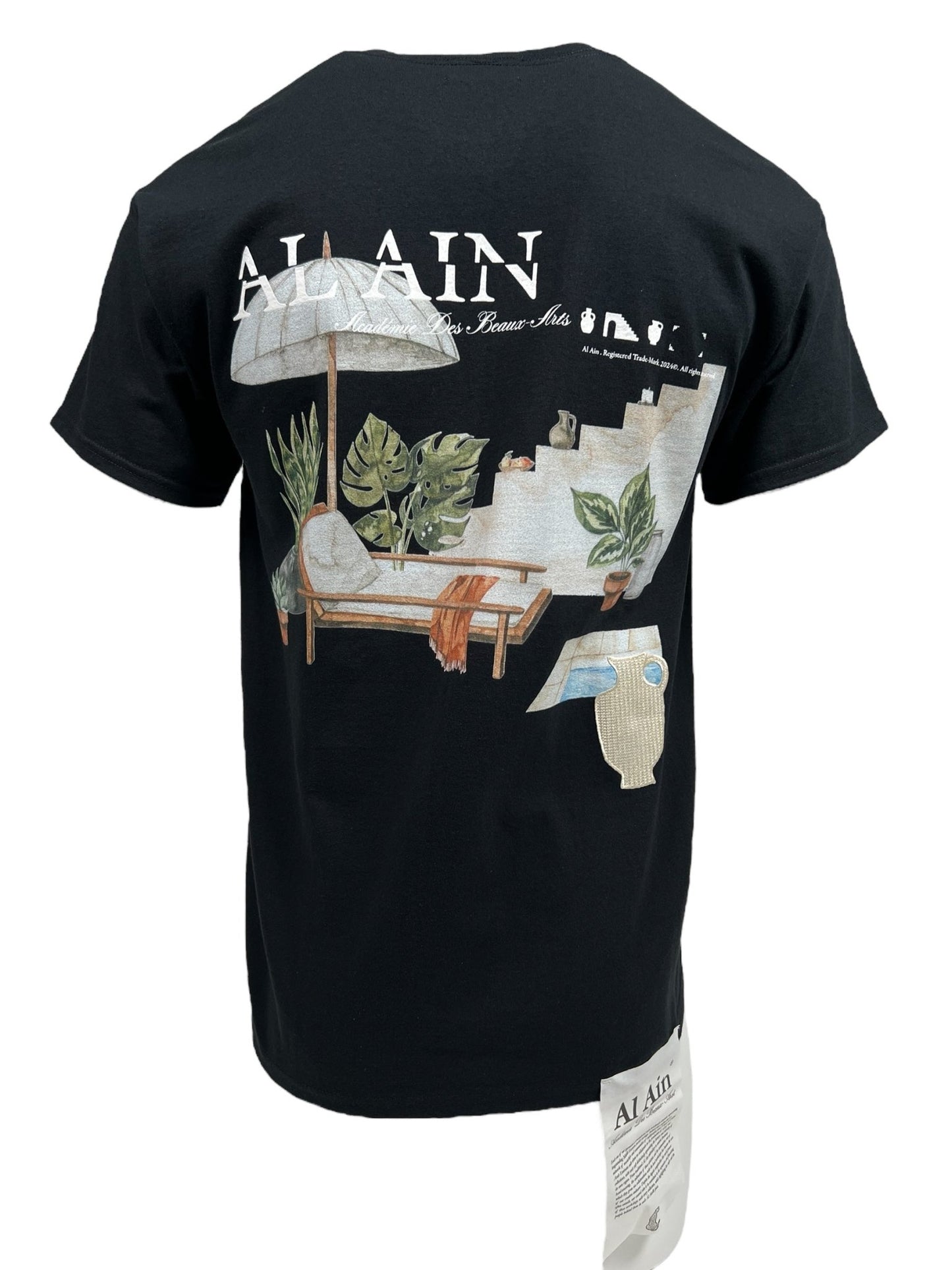 A black graphic T-shirt with a printed design on the back depicting a lounge chair, umbrella, and tropical plants. The text "AL AIN" is visible at the top. Made from 100% cotton, it features an attached tag at the bottom right. This product is the AL AIN AMHX S124 CHILL NOIR from AL AIN.