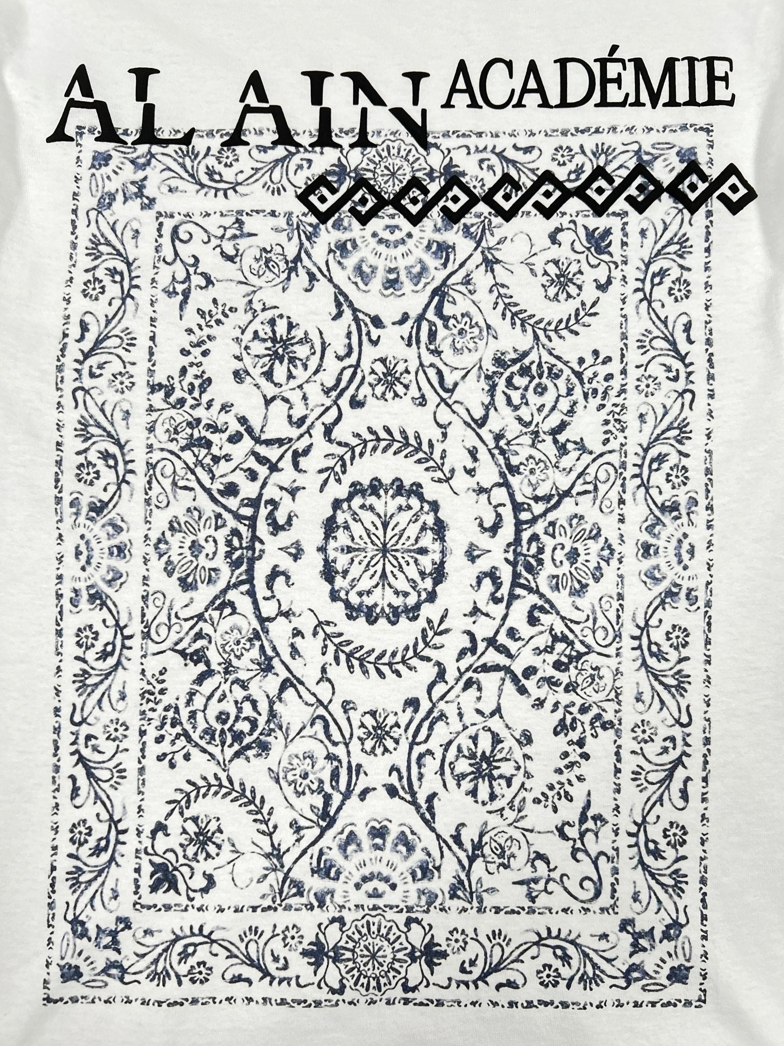 The Al Ain AMHX-S817 Sekaya T-shirt in white features a square ornate blue and white Persian-style floral rug print below the partially visible "ALAIN ACADÉMIE" text. Made of 100% cotton in Italy by AL AIN.