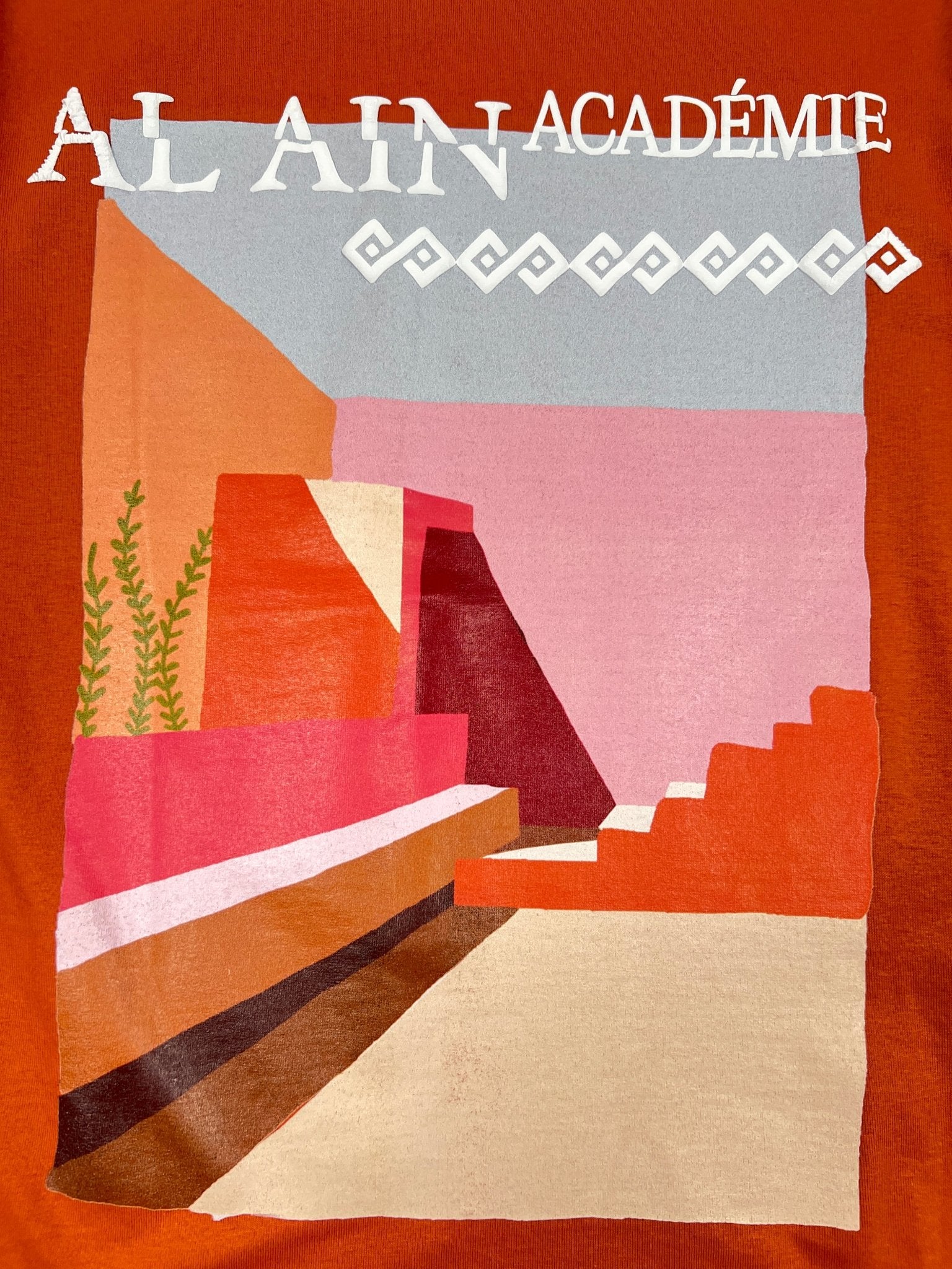 The AL AIN AMHX-S820 Almas T-shirt Ambra features abstract geometric shapes in red, pink, and beige on orange with "AL AIN ACADÉMIE" text and a zigzag detail—crafted from 100% cotton in Italy.