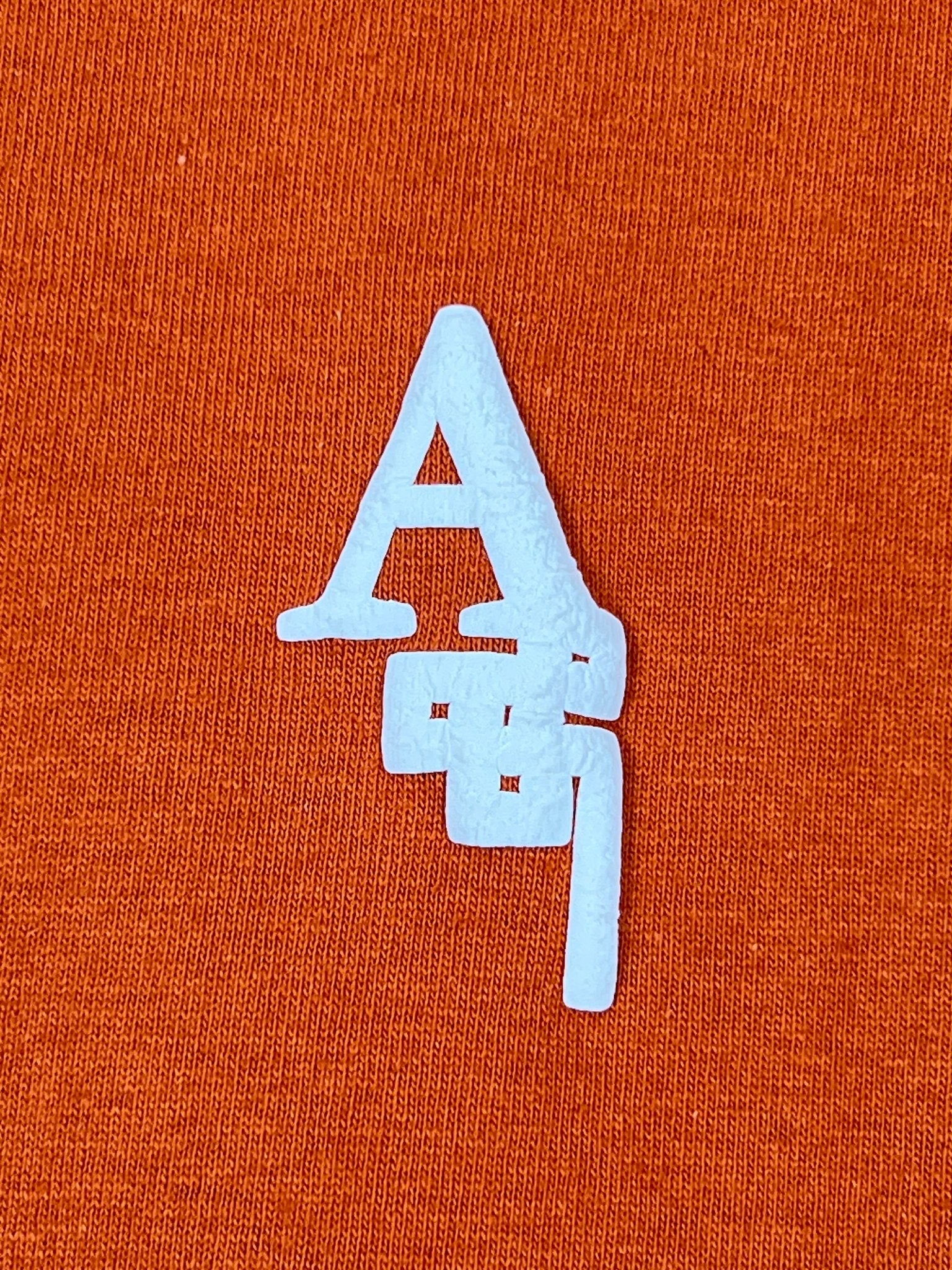 The AL AIN Al Ain AMHX-S820 Almas T-shirt Ambra features a white stylized "A" on orange 100% cotton, with kanji-inspired design elements; proudly made in Italy.