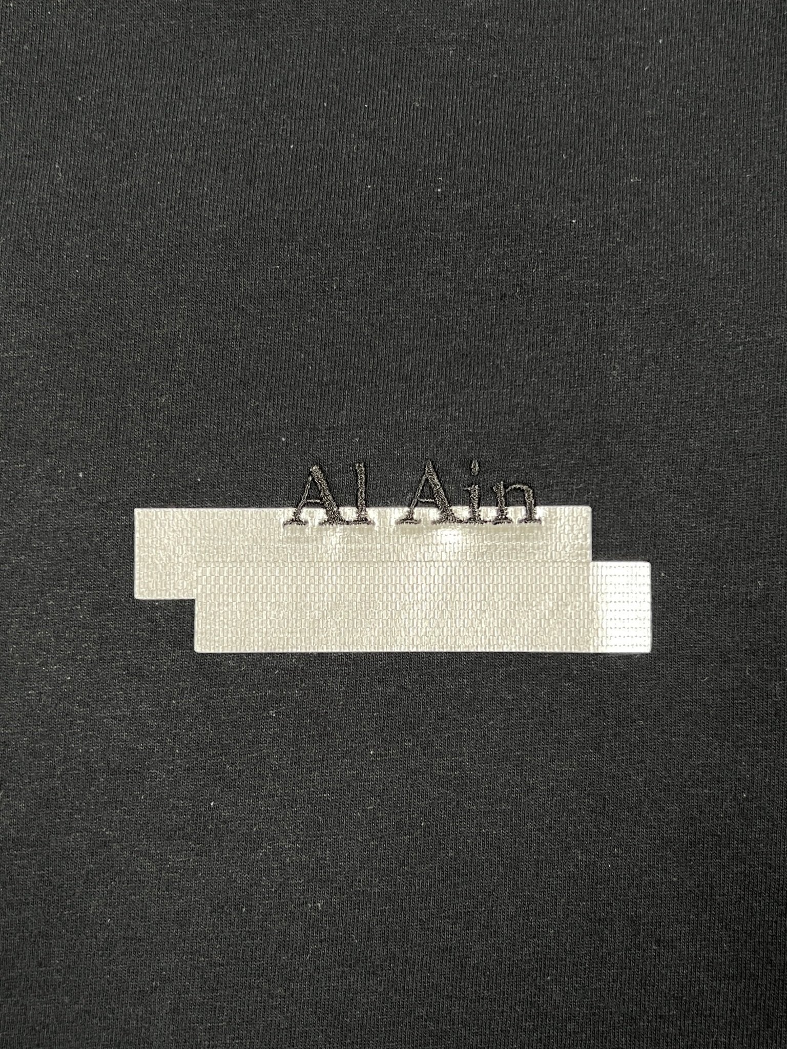 The AL AIN Al Ain AMHX-S870 Detruit T-shirt Black is a 100% cotton, Italian-made tee featuring "Al Ain" in silver above three overlapping rectangles in silver and white.