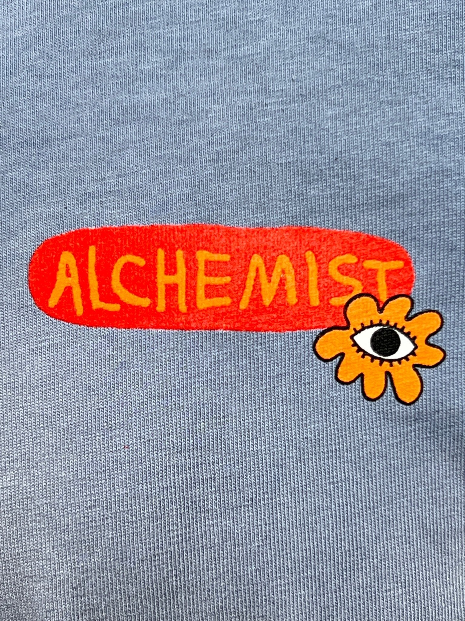 A vintage wash graphic t-shirt in gray displays "ALCHEMIST" in yellow letters on a red oval next to an orange flower with an eye. This design is featured on the Alchemist All-Seeing Cactus Tee Breeze Blue by ALCHEMIST.