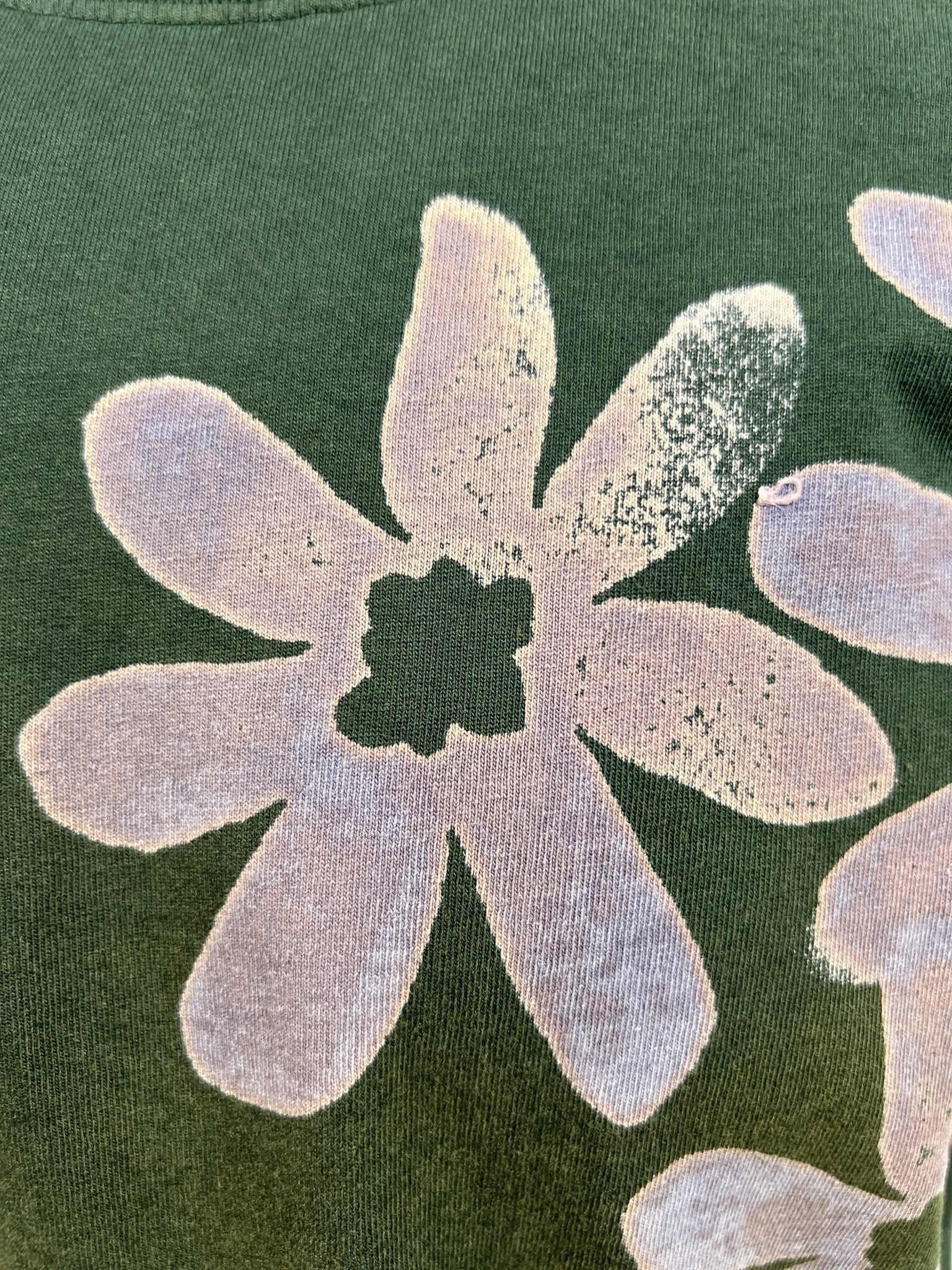A close-up of the ALCHEMIST BLOCK PRINT S/S TEE in moss green featuring a faded white flower design, showcasing the ALCHEMIST vintage washed finish. The flower displays a hole in the center and shows signs of wear.