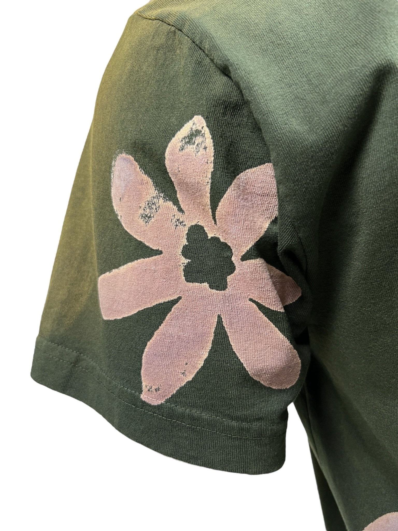 A close-up of the sleeve of the ALCHEMIST BLOCK PRINT S/S TEE in moss green, showcasing a pink floral graphic with a distressed look. The worn-away print enhances its vintage washed finish.