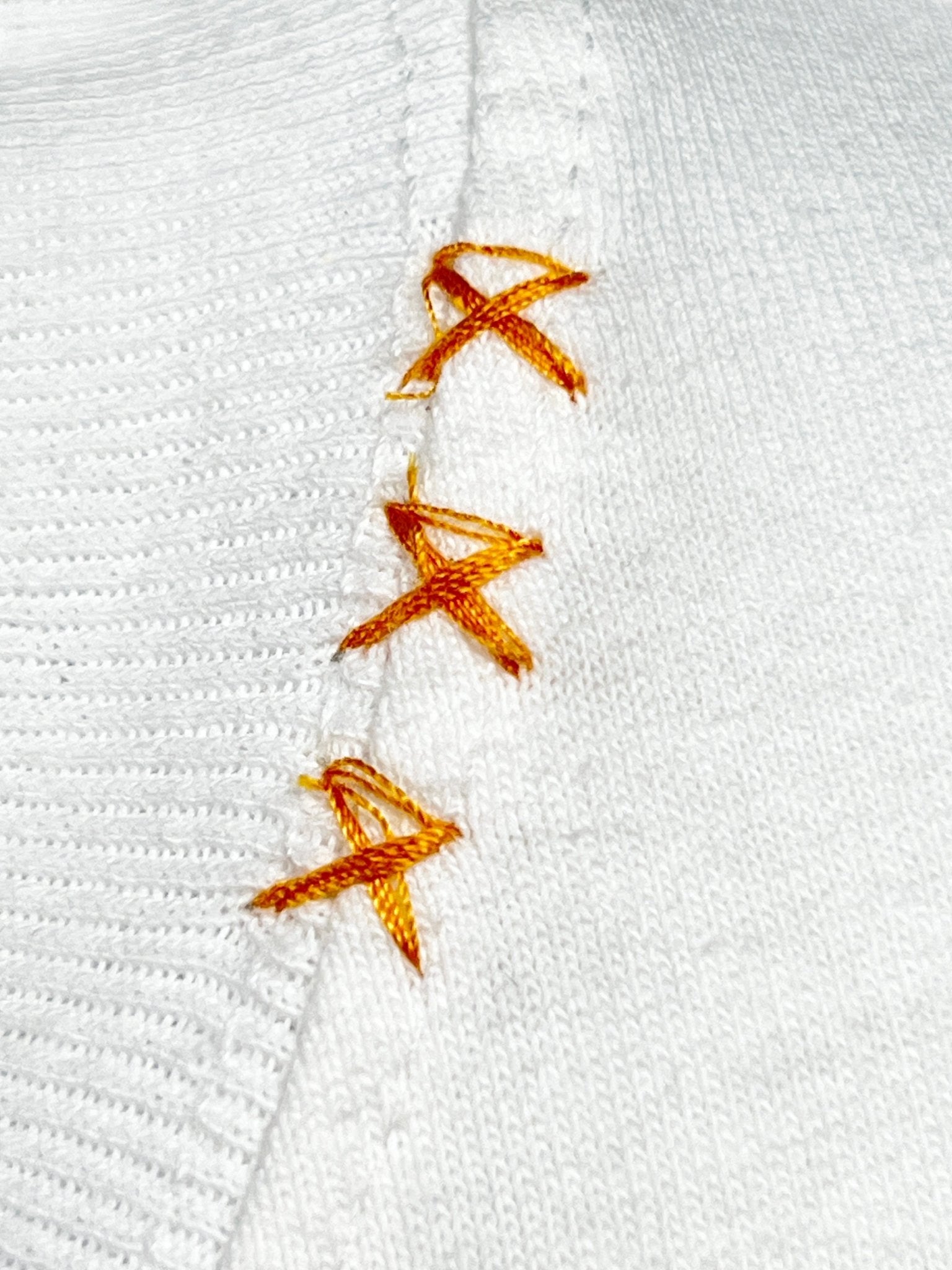 A close-up of a white fabric with orange cross-stitch embroidery along a seam, reminiscent of the intricate details found in an ALCHEMIST Alchemist Floral Skull Tee 2 White.
