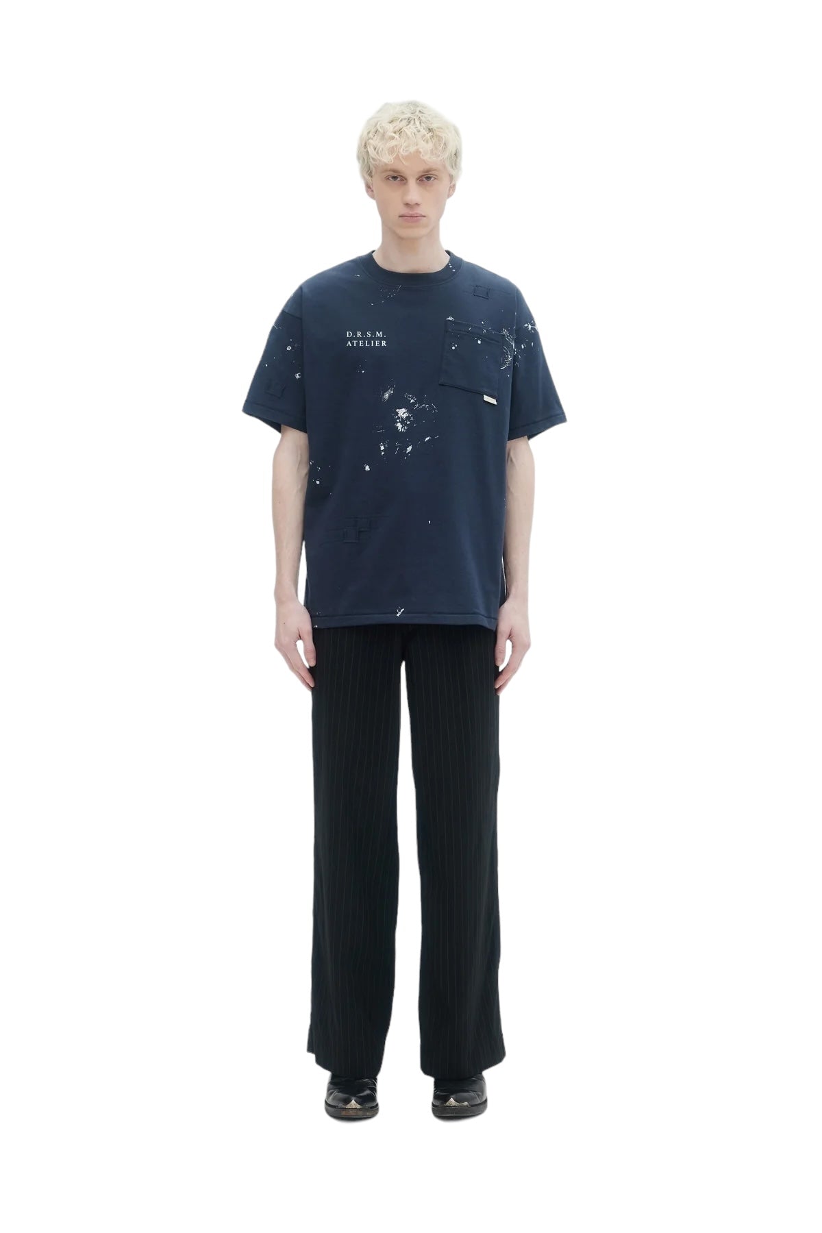 A person with short, light hair wears a dark blue Domrebel Atelier Pocket Tee by DOM REBEL and striped black pants, set against a white background.