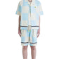 Against a plain backdrop, someone in sunglasses wears a BARROW multicolor checkered short-sleeve button-up shirt and matching Barrow S5BWUABE074 Popeline Shorts Vanilla in light blue and white cotton, complemented by black boots and socks.