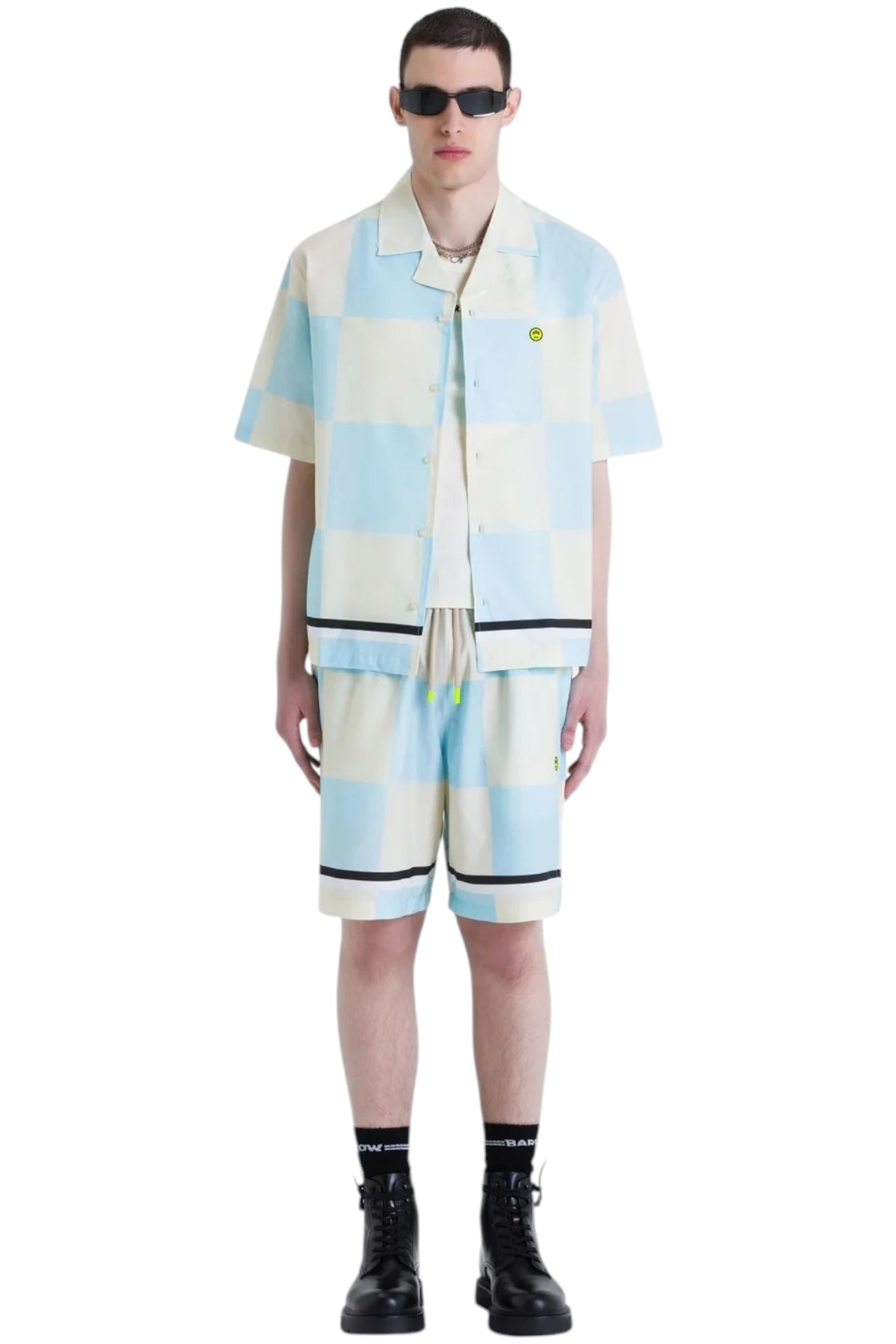 Against a plain backdrop, someone in sunglasses wears a BARROW multicolor checkered short-sleeve button-up shirt and matching Barrow S5BWUABE074 Popeline Shorts Vanilla in light blue and white cotton, complemented by black boots and socks.