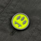 A round pin with a neon yellow-green background and black sad face is attached to black fabric, giving the BARROW S5BWUAJK065 Jacket Unisex Nero/Black an edgy urban accent perfect for accessorizing your look.